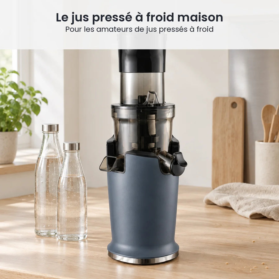 Extracteur de jus vertical bleu ardoise posé sur un plan de travail en bois clair, à côté de bouteilles en verre vides