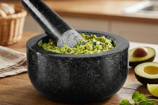 Guacamole maison dans un mortier en granit noir, le pilon posé dans la purée d'avocat, des demi-avocats et un quartier de citron vert à côté
